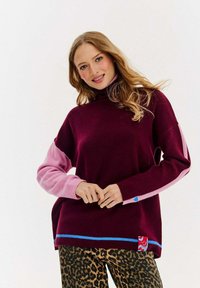 Burgundowy sweter z różowymi rękawami i niebieskimi akcentami. Posiada wysoki kołnierz i boczną metkę z logo, zestawiony z spodniami w panterkę.