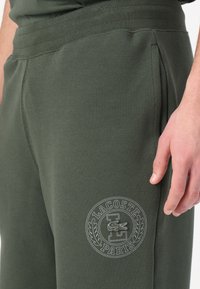 Pantalones de chándal verdes con una tela texturizada, que presentan un logo circular de Lacoste bordado en blanco en el muslo, cinturilla elástica y bolsillos laterales.