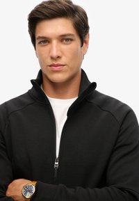 Schwarze Zip-Jacke mit hohem Kragen, strukturiertem Stoff und silbernen Reißverschlussakzenten. Darunter ein weißes Shirt und eine goldene Uhr am Handgelenk.