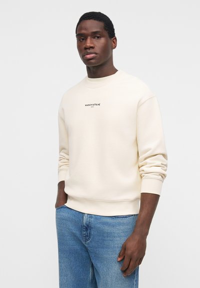 Νέος άνδρας με ανοιχτόχρωμο φούτερ Maison Kitsuné και μπλε τζιν, όρθιος με το ένα χέρι στην τσέπη, μπροστά από απλό φόντο.