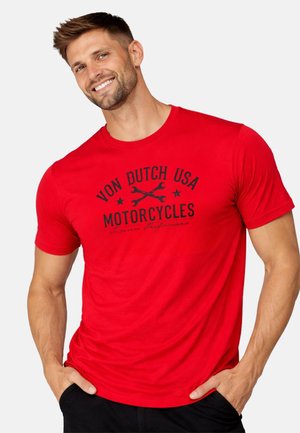 Homme souriant aux cheveux courts portant un T-shirt rouge avec l'inscription « Von Dutch USA Motorcycles » avec des icônes de clés croisées.