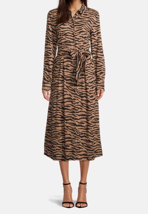 Frau trägt ein knielanges Kleid mit langen Ärmeln und Knöpfen, mit braunen und schwarzen Tigerstreifen und passendem Taillengürtel, kombiniert mit schwarzen Schuhen mit Knöchelriemen.