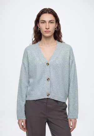 Femme aux cheveux bruns ondulés portant un cardigan gris clair boutonné et un pantalon gris foncé, debout devant un fond blanc uni.