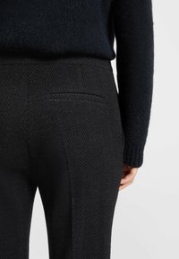 Mustat herringbone-kuvioiset housut, joissa on takatasku, yhdistettynä tumman, teksturoidun neuleen kanssa. Lähikuva vyötäröstä ja takaosasta.