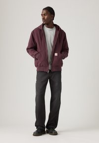 Μπουφάν με κουκούλα σε μπορντό χρώμα και φερμουάρ, γκρι πουκάμισο από κάτω, συνδυασμένο με σκούρο παντελόνι και μαύρα παπούτσια. Απλός σχεδιασμός, χωρίς έντονα μοτίβα.