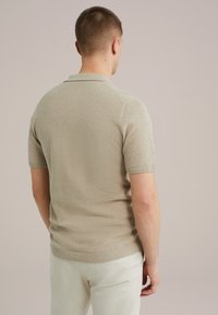 Bež majica s kratkimi rokavi iz pletenine s teksturiranim vzorcem, z ribastim ovratnikom in manšetami, v kombinaciji s svetlimi hlačami.