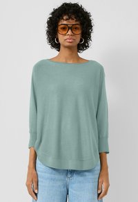 Kobieta z kręconymi włosami nosi dużą parę amber okular�ów przeciwsłonecznych, srebrne kolczyki w kształcie kół, luźny sweter w jasnym odcieniu turkusu oraz jasnoniebieskie dżinsy na jednolitym tle.