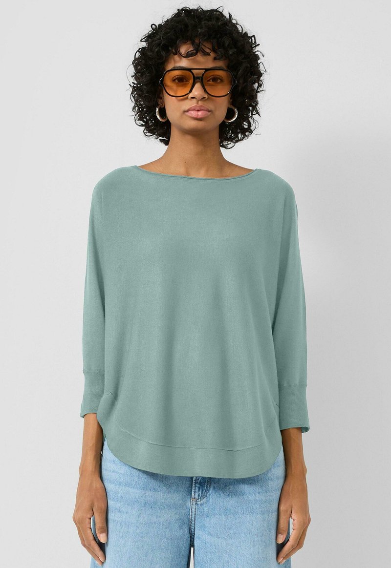 Kobieta z kręconymi włosami nosi dużą parę amber okularów przeciwsłonecznych, srebrne kolczyki w kształcie kół, luźny sweter w jasnym odcieniu turkusu oraz jasnoniebieskie dżinsy na jednolitym tle.