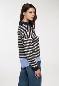 Pullover a righe in blu navy, crema e azzurro chiaro con polsini a coste. Presenta un colletto e un effetto sovrapposto con una camicia blu sotto.