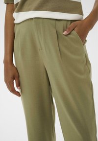 Pantalon vert tailleur en tissu doux, avec une texture lisse, des plis et des poches latérales. Une coupe ajustée avec un design à jambe droite.