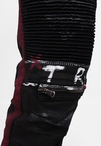 Jeans neri e bordeaux con effetto usurato, caratterizzati da costine testurizzate, una piccola tasca con cerniera e audaci dettagli in vernice bianca e rossa.