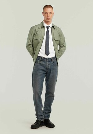 Uomo che indossa una giacca verde con zip sopra una camicia bianca con cravatta scura, jeans blu e scarpe nere, in piedi davanti a uno sfondo chiaro e semplice.