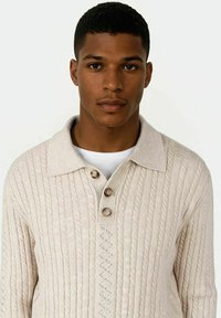 Beige strikket genser med ribbet tekstur, med krage og tre knapper. Enkel, uformell design med subtile mønsterdetaljer.