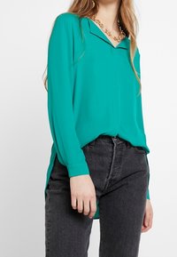 Blusa lunga teal con colletto a disfasso, spacchi laterali e vestibilità morbida, abbinata a jeans grigi scuri a vita alta. Collana metallica visibile.