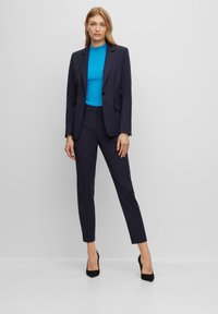Námořnický oblek na míru se sako na jedno knoflík, slim-fit kalhoty a světlý modrý žebrovaný top. Černé vysoké podpatky dotvářejí celý outfit.