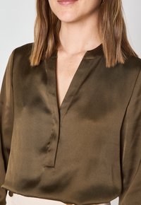 Blusa di raso verde oliva con scollo a V, maniche lunghe e leggero lucido. Presenta un design minimale e una texture morbida.