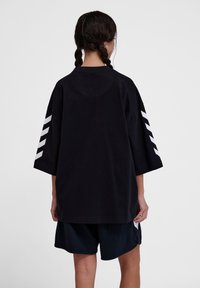 Zwart oversized t-shirt met een ronde halslijn en korte mouwen, voorzien van witte hoekige strepen op de mouwen. Draagt met donkere shorts.