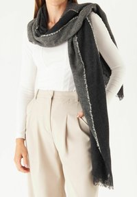 Écharpe noire et grise avec des bords effilochés, fabriquée dans un matériau doux, drapée de façon décontractée. Elle présente un motif texturé et complète un haut blanc.