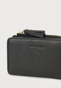 Musta teksturoitu nahkalompakko, jossa on kultainen vetoketju ja tupsun vetoketjuvetäjä, sekä kohokuvioitu brändinimi "Coccinelle" etupuolella.