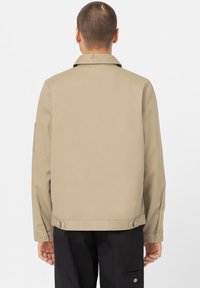 Man sett bakifrån iförd en beige jacka med knäppte ärmar och svarta byxor med en liten logotyp på fickan.