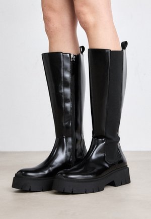 Bottes noires brillantes montantes jusqu'au genou avec des semelles épaisses à crampons, portées sur des jambes nues, dotées d'une fermeture éclair latérale et de languettes en haut.