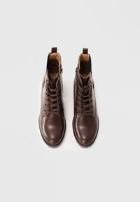 Bottines en cuir marron avec fermeture à lacets, orteils ronds et zips latéraux. Présentent une texture lisse et des semelles en caoutchouc noir contrastantes.