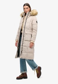 Polstret beige frakke med faux fur hætte, frontlynlås, flere lommer, parret med denim jeans med optrullede manchetter og brune snørebåndsstøvler.