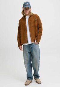 Braunes Cordhemd über einem weißen T-Shirt, blaue Jeans und beige Schuhe. Eine blaue Mütze mit Text rundet das Outfit ab.