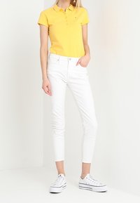 Polo shirt jaune avec un col, associé à un pantalon blanc slim et des baskets blanches. La coupe est ajustée et le tissu semble lisse.