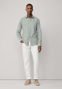 Camicia verde a quadretti con maniche lunghe, collo alla francese, abbinata a pantaloni bianchi e scarpe beige, in piedi su uno sfondo neutro.