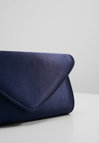 Pochette enveloppe en satin bleu foncé avec un rabat triangulaire, reposant sur une surface blanche sur un fond gris clair uni.