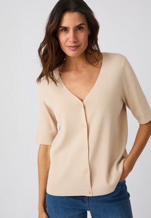 Donna con capelli scuri che indossa una maglietta beige a maniche corte con bottoni e jeans blu, in piedi con una mano in tasca davanti a uno sfondo semplice.
