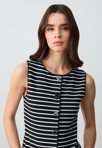 Gestreiftes ärmelloses Kleid mit schwarz-weißen horizontalen Streifen, rundem Ausschnitt und dekorativen silbernen Knopfakzenten auf der Vorderseite.