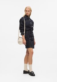 Robe chemise en denim bleu foncé avec des coutures contrastées, manches longues, fermeture à boutons et une coupe ajustée. Accessoirisée avec un sac rond.