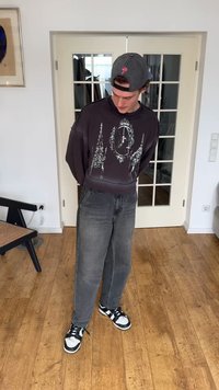 Noor mees toas, kandes tagurpidi halli mütsi, musta dressipluusi valge graafikaga, hallid teksad ja must-valged tossud, seistes puitpõrandal.