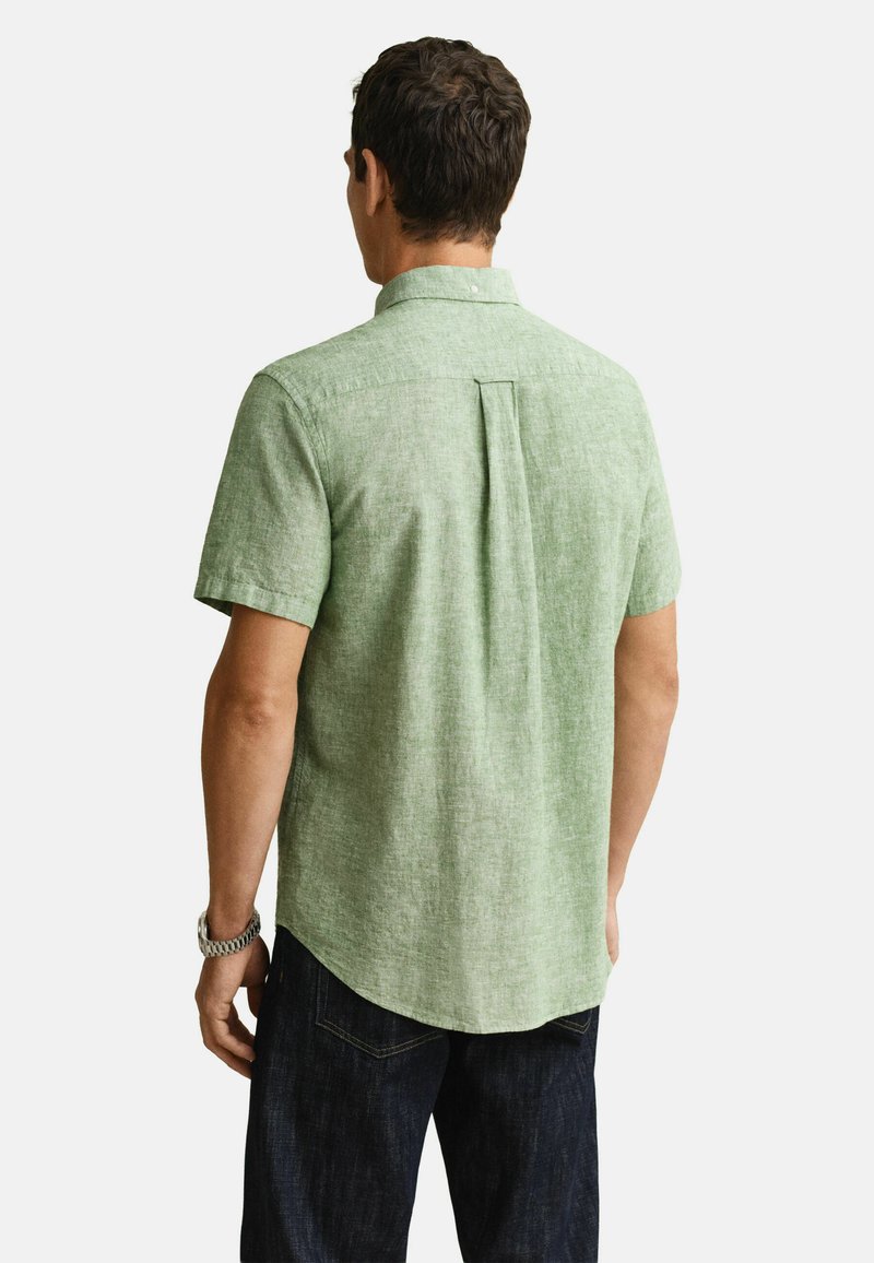 Mand iført en lysegrøn kortærmet linskjorte med en bagpå bokspliséring, kombineret med mørkeblå jeans og et sølvfarvet armbåndsur.