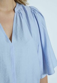 Blouse bleu clair avec un col en V et des manches courtes bouffantes. Le tissu semble texturé avec de légères rayures. Dispose d'une fermeture à boutons sur le devant.