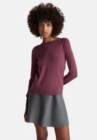 Mladá žena s kudrnatými vlasy, která má na sobě fialový top s dlouhými rukávy a odhalenými rameny a šedou sukni, pózující před světlým bílým pozadím.