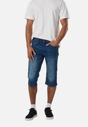 Homme portant un t-shirt blanc, un short en jean bleu au niveau des genoux, des chaussettes blanches et des baskets noires, une main dans la poche sur fond uni.