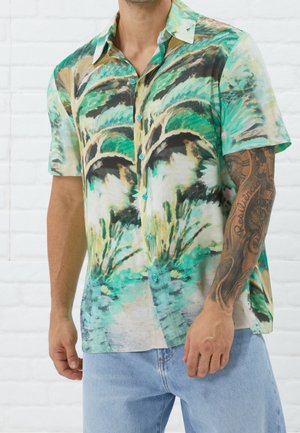 Mand iført kortærmet knap-skjorte med grøn og beige abstrakt naturmønster og lyseblå jeans, der viser tatoveret højre arm.