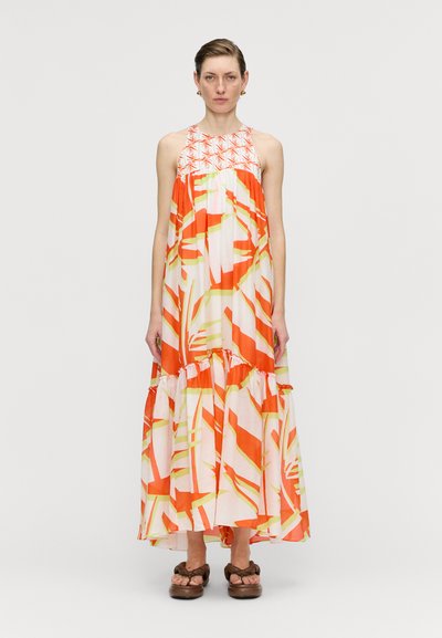 Vestido maxi de tejido naranja y blanco con patrones de hojas verdes, con escote halter y dobladillo acampanado en capas. Llevado con zapatos marrones.