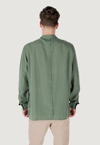 Maglietta verde a maniche lunghe con una texture liscia, dettaglio della cucitura sul retro e chiusura con bottoni ai polsini. Accompagnata da pantaloni di colore chiaro.