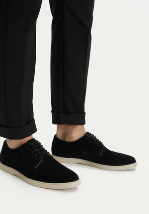 Scarpe stringate in suede nero con suola bianca indossate con pantaloni neri risvoltati su sfondo chiaro.