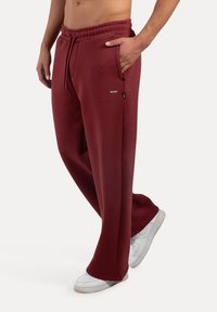 Pantalones deportivos burdeos de tejido suave con cintura elástica, bolsillos laterales y una pequeña etiqueta de marca cerca del bolsillo. Llevados con zapatillas blancas.