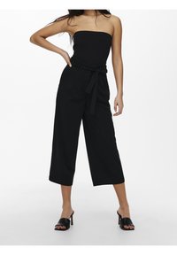 Top noir sans bretelles associé à un pantalon noir taille haute à jambes larges et des talons glissés noirs. Texture lisse, design minimaliste, sans motifs.
