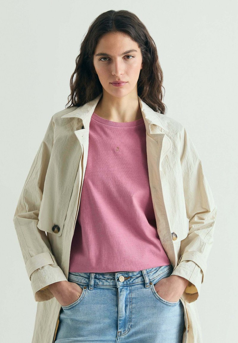 Tânără cu păr lung și întunecat poartă o jachetă bej peste un tricou roz și blugi bleu deschis, stând cu mâinile în buzunare.