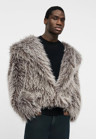 Νέος άνδρας, φοράει γκρι χνουδωτό πλαστικό γούνινο μπουφάν πάνω από μαύρο πουκάμισο, στέκεται με τα χέρια στις τσέπες μπροστά σε απλό λευκό φόντο.