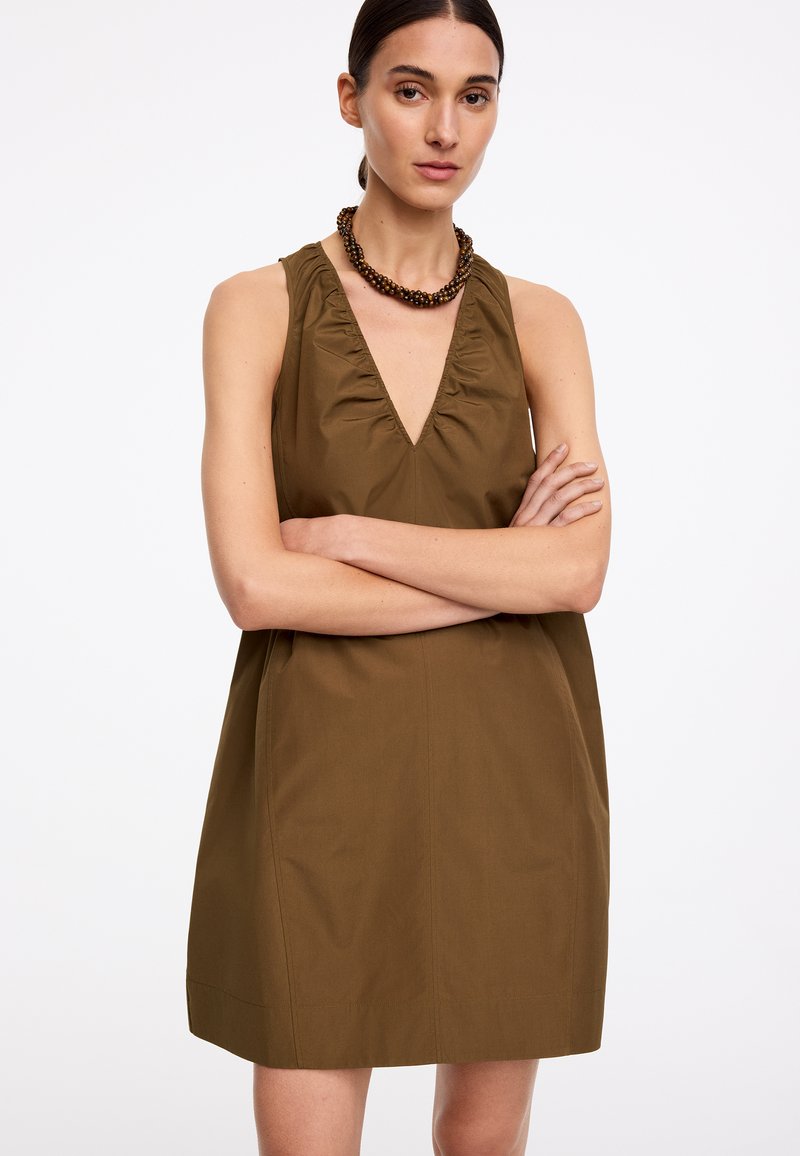 Femme portant une robe sans manches vert olive à décolleté en V profond et un collier de perles, debout les bras croisés.