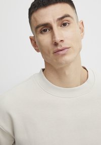 Sudadera beige clara con cuello redondo y tejido texturizado, que cuenta con una superficie suave y puños acanalados, sobre un fondo blanco.