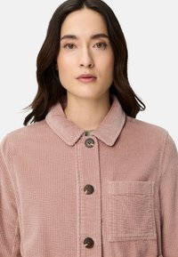 Różowa sztruksowa kurtka z kołnierzem, pięcioma guzikami i kieszenią na piersi. Posiada teksturowany, prążkowany wzór.